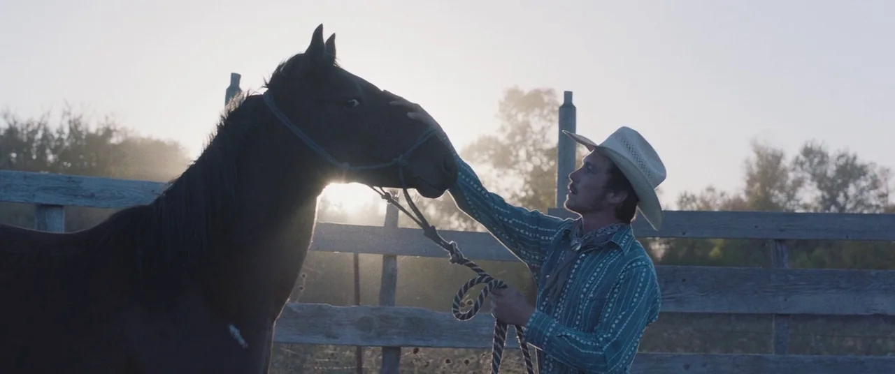 The Rider (2017) movie still: medium — A man stands in a wooden corral during the golden hour of the day. He strokes the forehead of a dark horse…