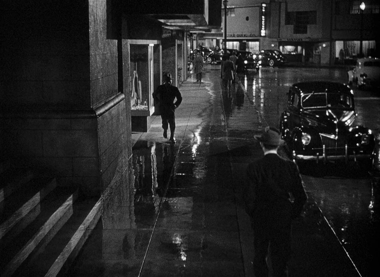 Dead Reckoning (1946) movie still: wide — A rain-drenched urban sidewalk at night features reflective puddles and storefront windows. Several men in…