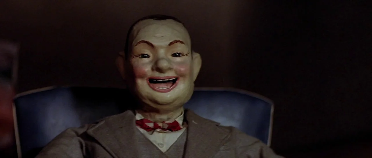 The Funhouse (1981) movie still: closeup — A dimly lit room holds a wooden ventriloquist dummy sitting upright. The dummy remains perfectly still…