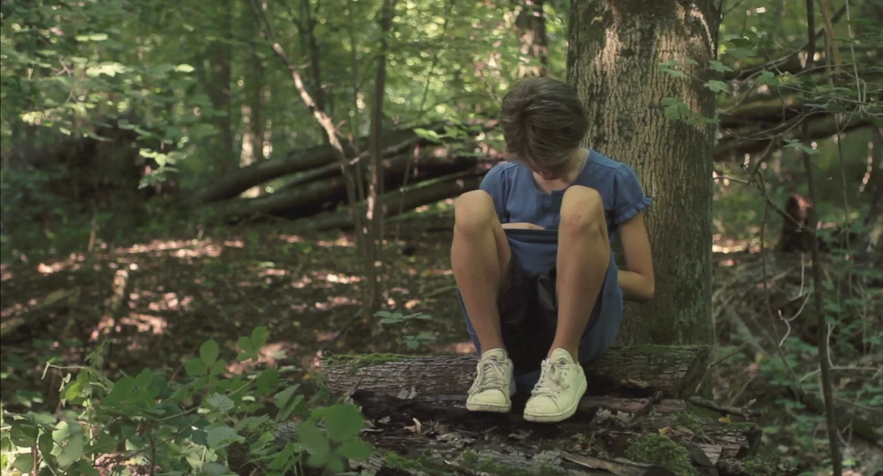 Tomboy (2011) movie still: medium — A dense forest floor with moss-covered logs and scattered foliage under a tree canopy. A young girl sits on a…
