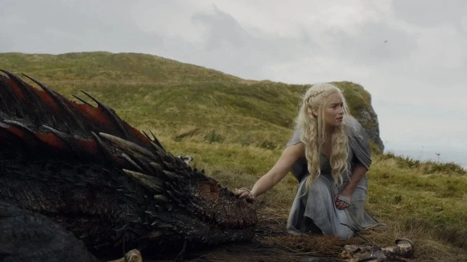 Game of Thrones (2015) movie still: medium — A woman in a grey gown kneels on a grassy cliffside next to a dragon. The woman reaches out to touch…