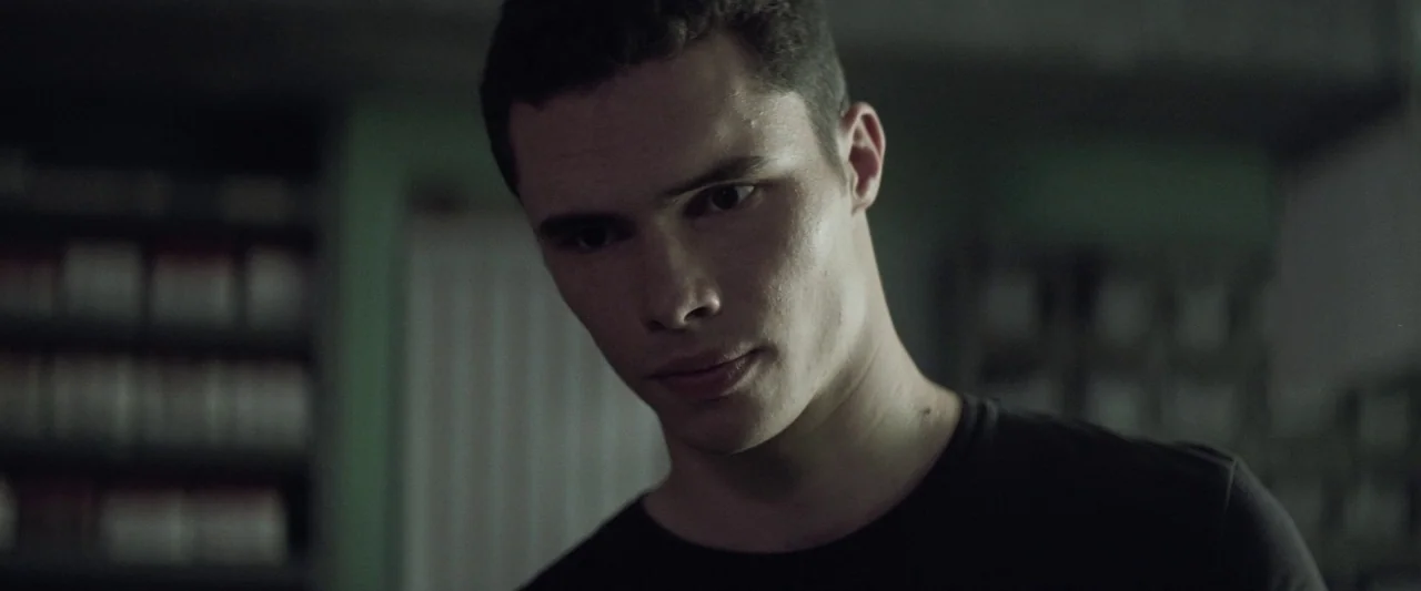 Colt 45 (2014) movie still: closeup — A dark, dimly lit office contains rows of shelves filled with white binders. A young man with short dark hair…