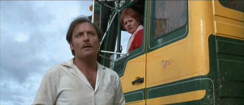 Roadgames (1981) movie still: medium — A man and a woman stand next to a large yellow and green truck. The sky is overcast and cloudy above the…