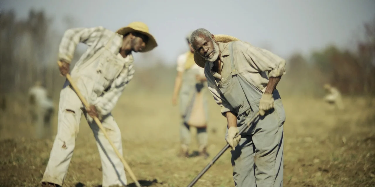 Green Book (2018) movie still: medium — An open rural field with dry, sparse vegetation and light dust in the air. Several older men in worn work…