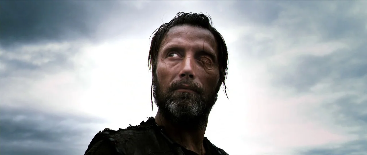 Valhalla Rising (2009) movie still: closeup — A man with a scarred face stands under a heavily overcast grey sky. He looks towards the side while…