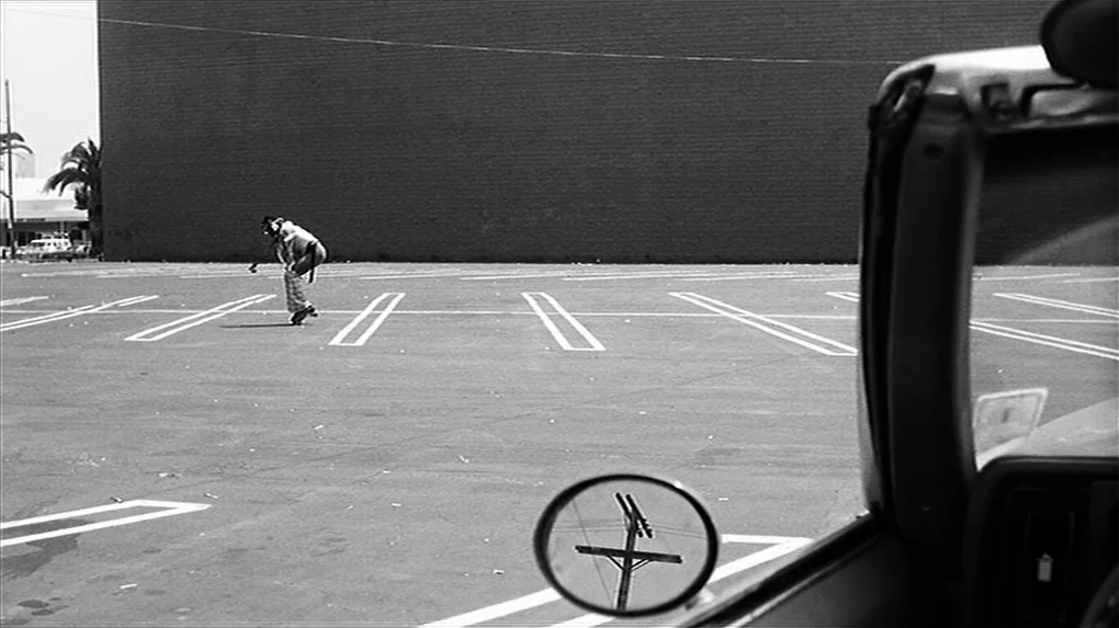 The State of Things (1982) movie still: wide — An empty asphalt lot features a large dark wall as a backdrop. A single person rolls across the space…