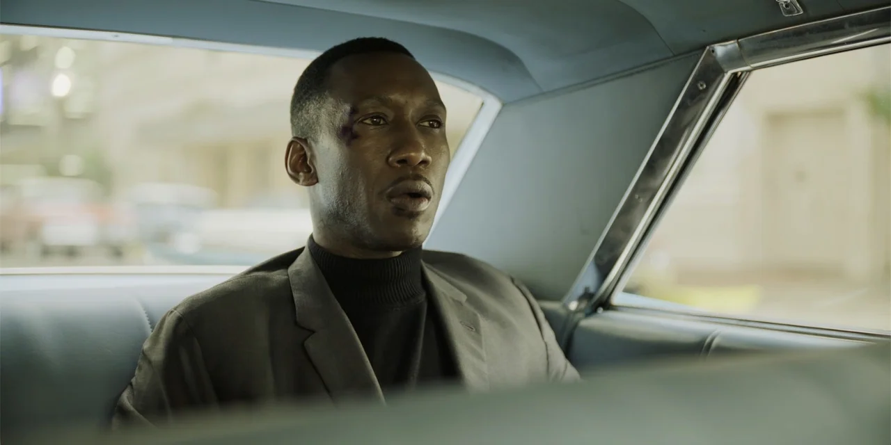 Green Book (2018) movie still: medium — A man sits in the blue leather backseat of a vintage car during the day. The man looks out the side window…