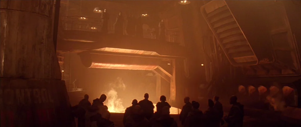 Alien 3 (1992) movie still: wide — A dark, cavernous industrial interior is filled with orange haze and structural metal supports. A crowd of people…