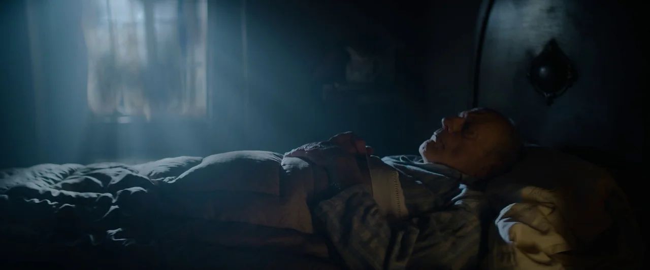 Nandor Fodor and the Talking Mongoose (2023) movie still: medium — A dark, sparsely lit room contains a bed with a wooden headboard. An elderly man…