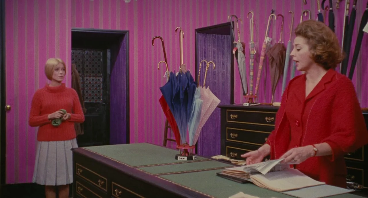 The Umbrellas Of Cherbourg (1964) movie still: medium — An interior space features bright magenta striped wallpaper and rows of hanging umbrellas. A…