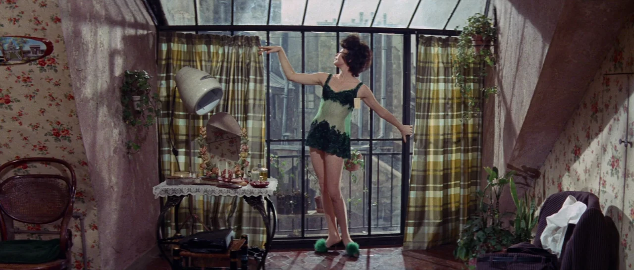 Irma La Douce (1963) movie still: fullbody — An attic room features floral wallpaper, a vanity table, and large sloping glass windows. A woman in a…