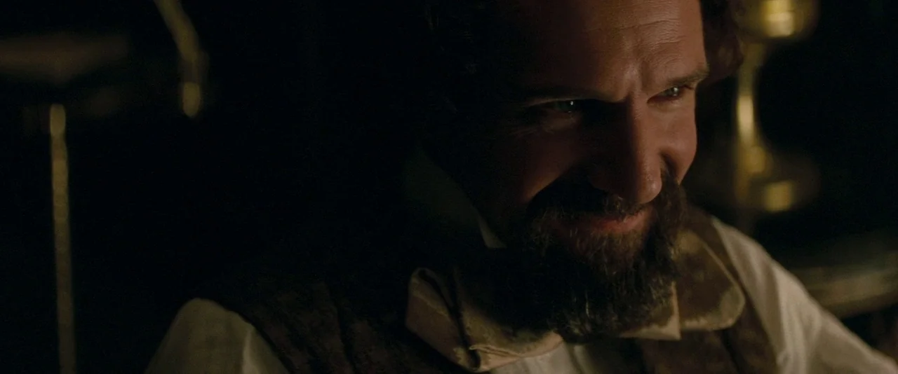 The Invisible Woman (2013) movie still: closeup — A dark, wood-paneled room is lit by a single warm practical lamp at night. A middle-aged man with…