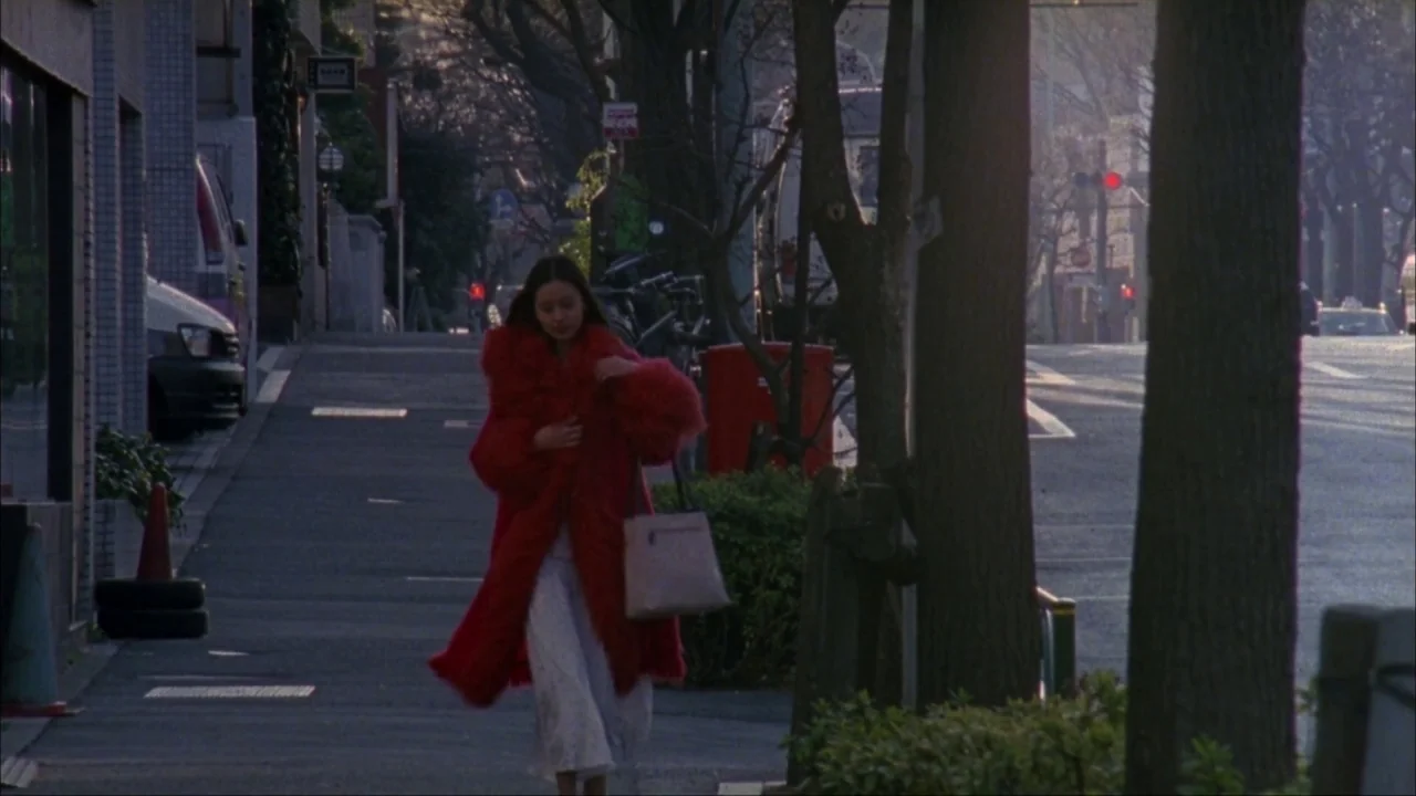 Audition (1999) movie still: fullbody — An urban sidewalk runs parallel to a road with parked cars and mature trees. A woman in a red coat walks…