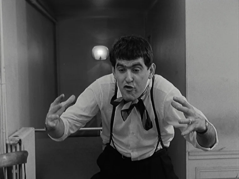 Vivre Sa Vie (1962) movie still: medium — A dimly lit hallway with a wall-mounted light fixture in the background. A man stands facing the camera…