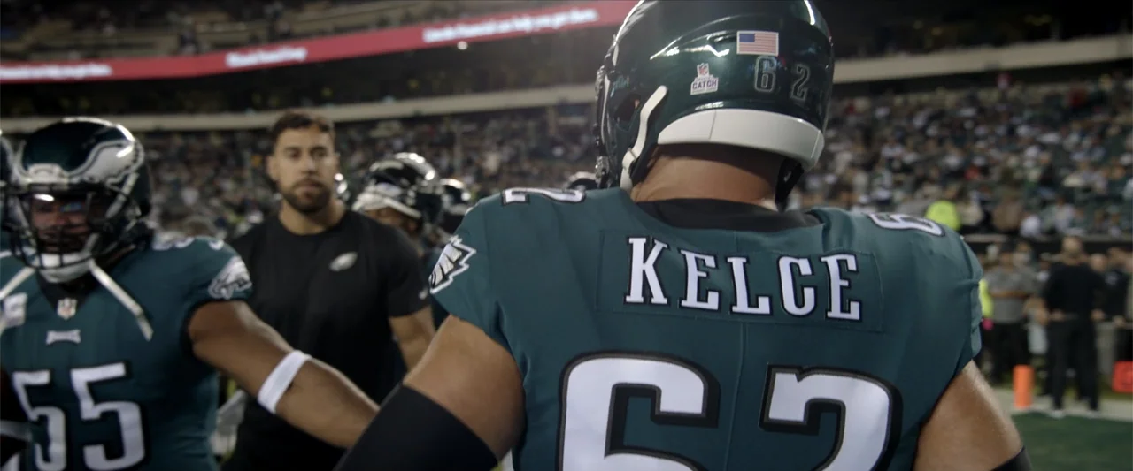 Kelce (2023) movie still: medium — A professional football stadium at night with players gathered on the sidelines. A player wearing jersey number…