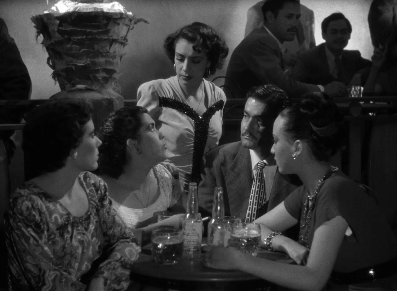 Victims of Sin (1951) movie still: medium — A dimly lit nightclub features a group of people sitting at a small table. A man in a suit engages in…