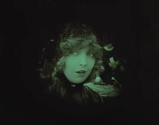 Birth of a Nation (1915) movie still: closeup — A dark parlor setting with figures barely visible in the surrounding deep shadow. A woman with short…