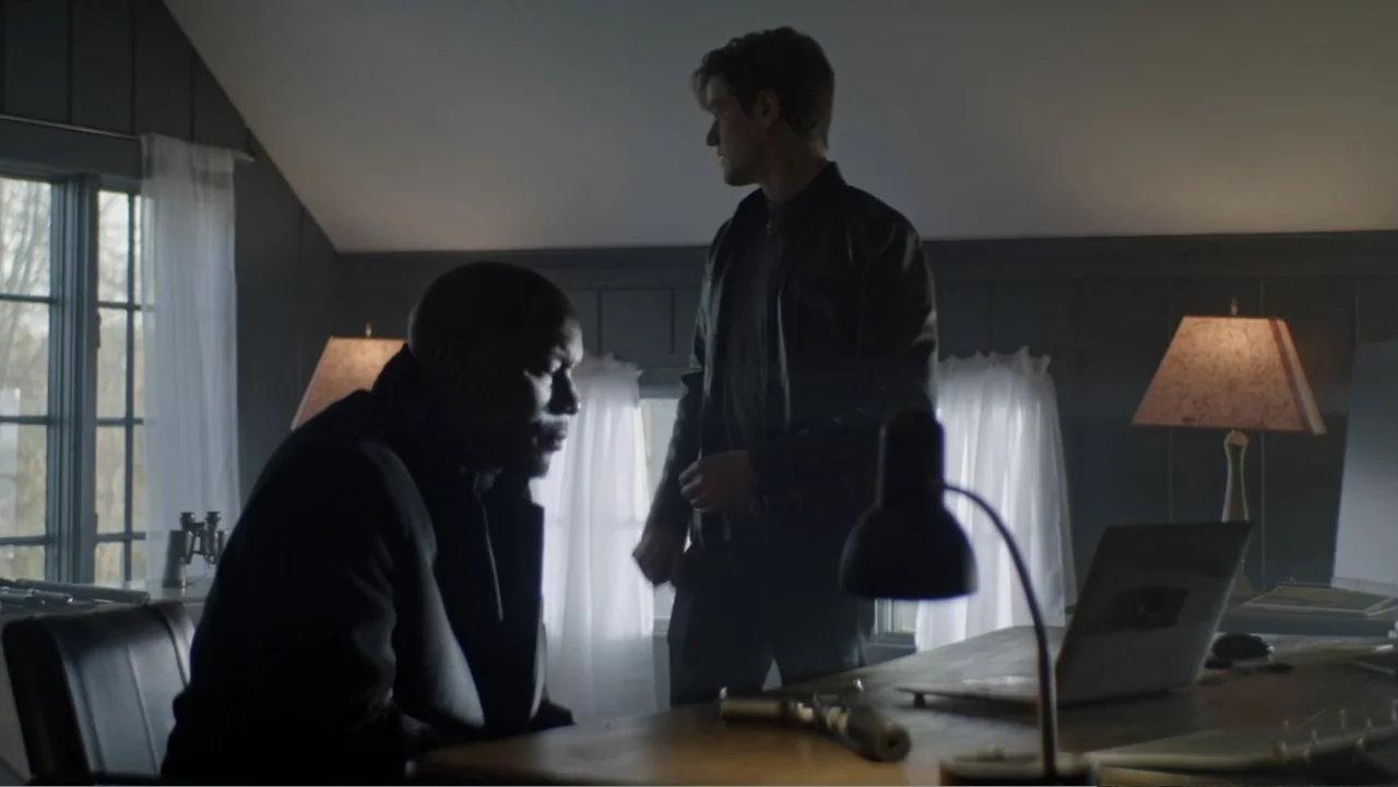 The Collective (2023) movie still: medium — A dark home office features a wooden desk with a laptop and a pistol. Two men are positioned in the…
