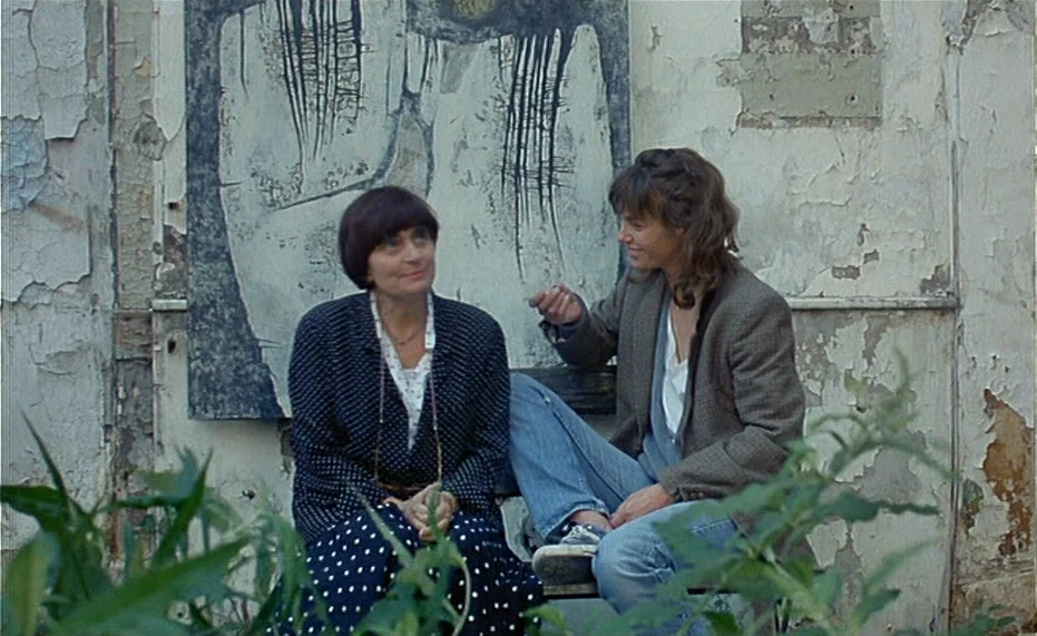 Jane B. par Agnès V. (1988) movie still: medium — A worn outdoor wall with peeling white paint sits behind two seated women. One woman wears a polka…