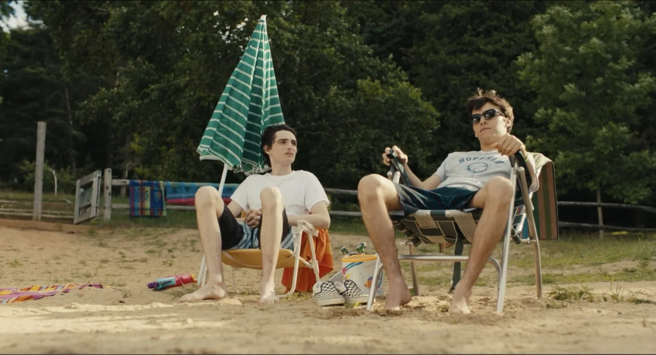 Hell of a Summer (2025) movie still: fullbody — Two teenage boys sit in folding chairs on a sandy expanse near trees. They are lounging in the…