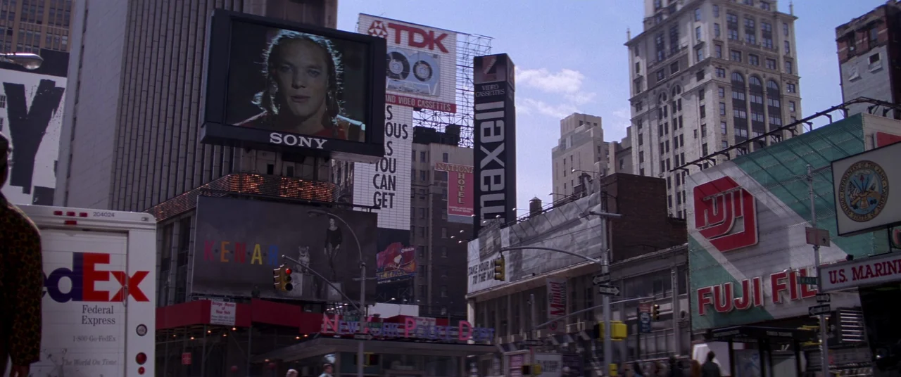 Hackers (1995) movie still: wide — A dense urban intersection features multiple large billboards for commercial brands and public services. The…