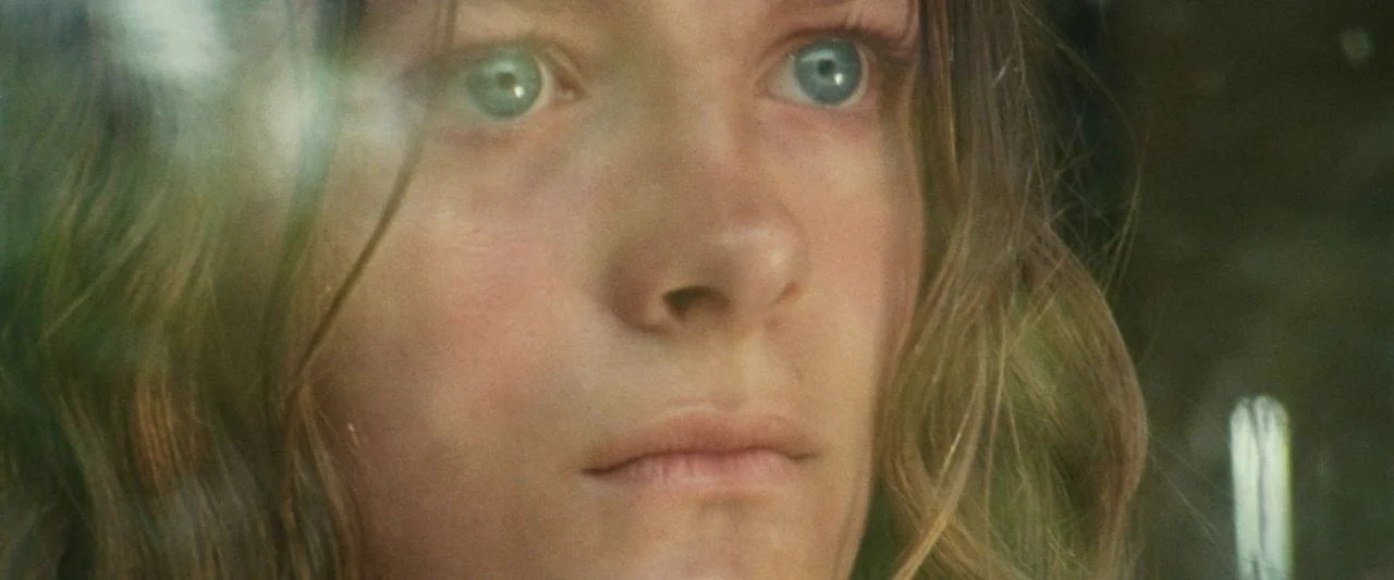 Adoration (2019) movie still: closeup — A close-up view of a young woman's face positioned behind a transparent surface. She gazes forward with…
