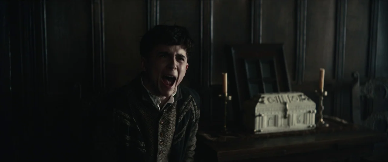 The King movie still: medium — A dimly lit room features dark wood-paneled walls and a small ornate white box. A young man sits in the foreground…