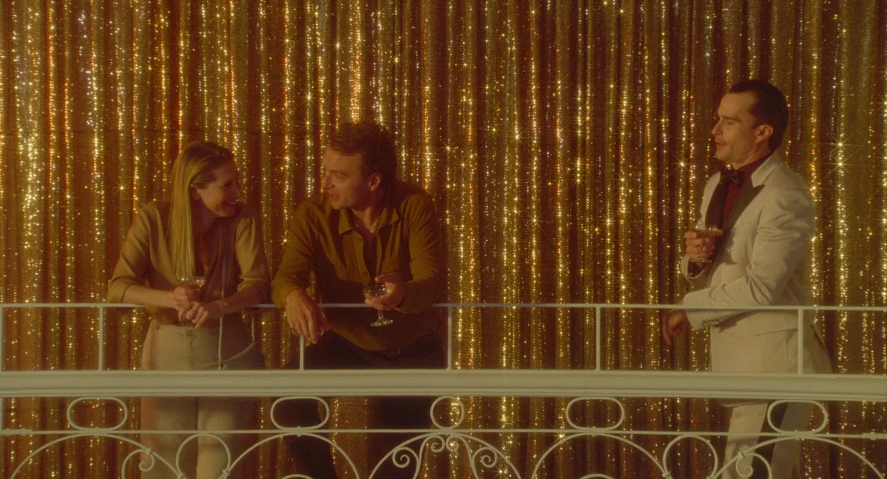 Babysitter (2022) movie still: medium — A festive balcony area features a decorative white metal railing against shimmering gold curtains. Two…