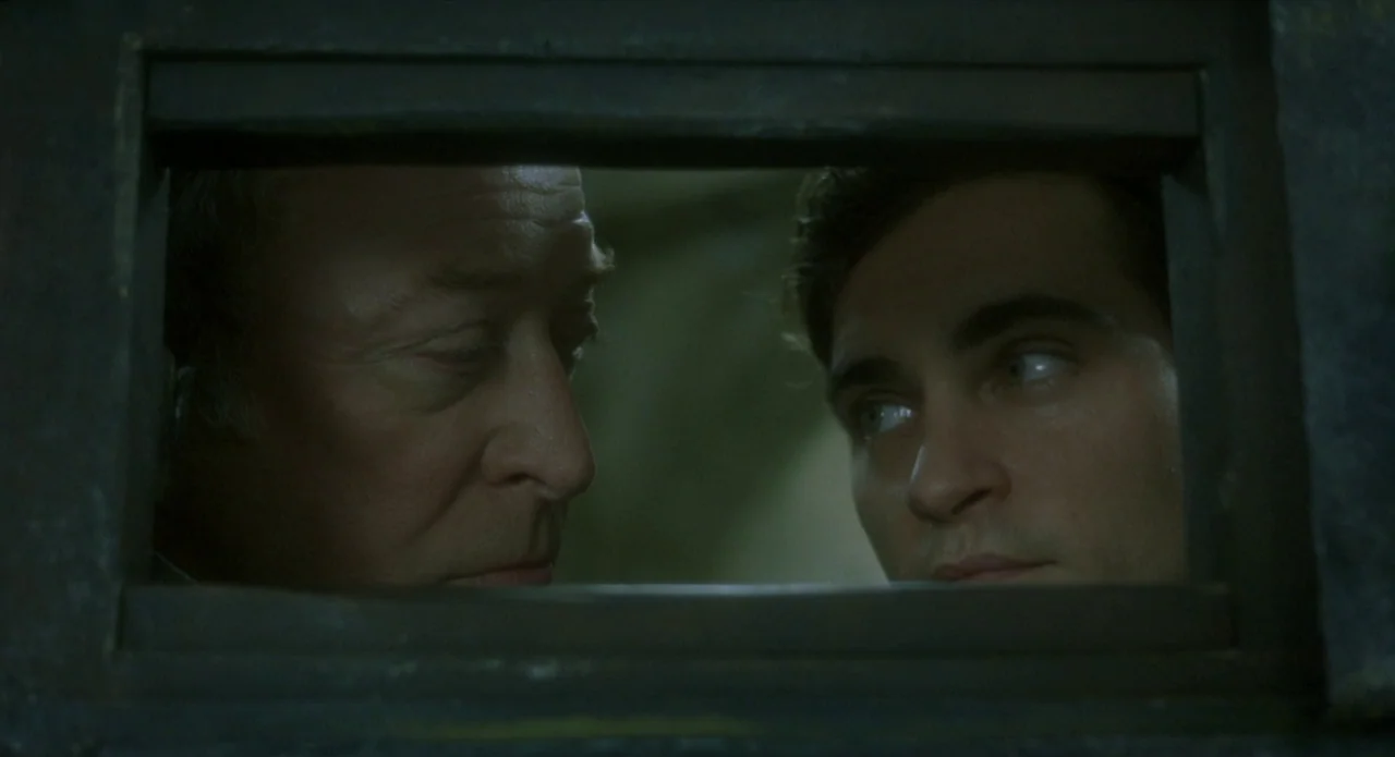 Quills (2000) movie still: closeup — Two men appear side-by-side behind a narrow, dark rectangular frame. The scene is lit to focus on their facial…