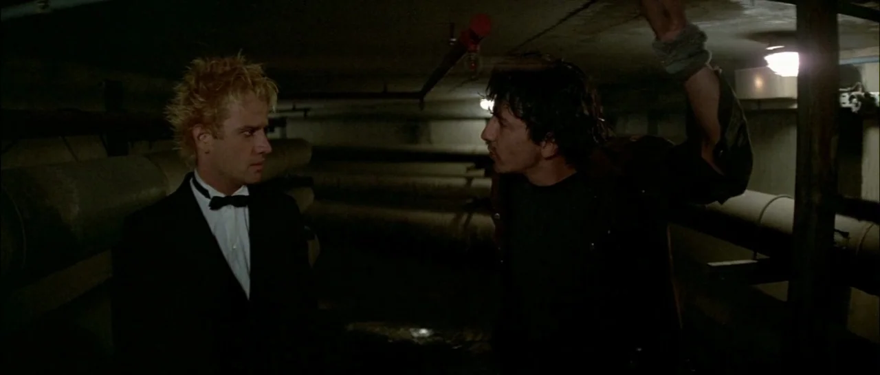 Subway (1985) movie still: medium — A subterranean area filled with large industrial pipes and dim overhead lighting. Two men face each other during…