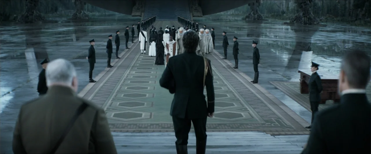 Dune (2021) movie still: wide — A vast exterior landing pad with a large ship looming above a concrete surface. A military officer walks toward a…