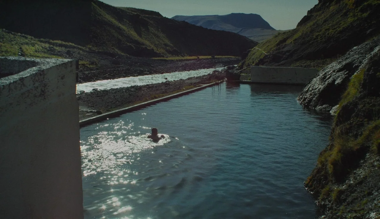 Land Ho! (2014) movie still: wide — An outdoor concrete hot spring pool is located in a deep mountain valley. A person is swimming in the water…