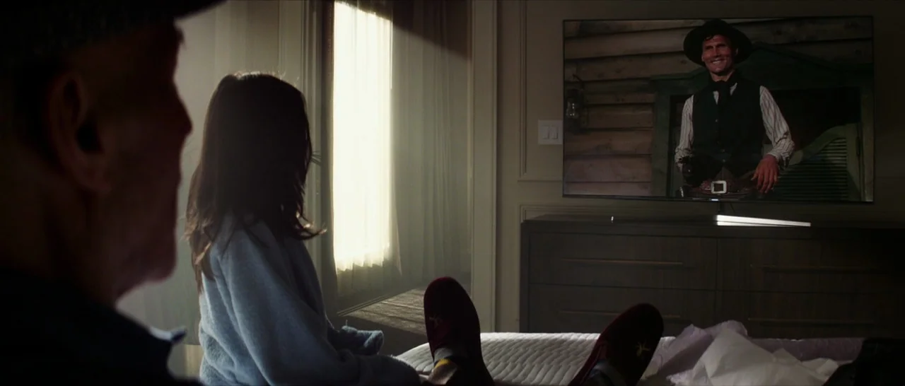 Logan (2017) movie still: medium — A dimly lit bedroom features a modern dresser supporting a large flat screen television. Two people are seated on…