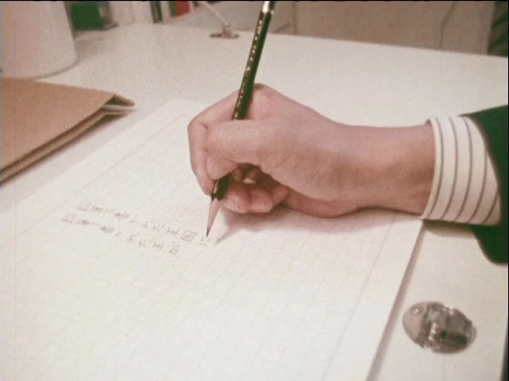 Catalogue of Memory (1977) movie still: closeup — A clean white desk features a grid-lined sheet of paper for manual handwriting. A hand in a…