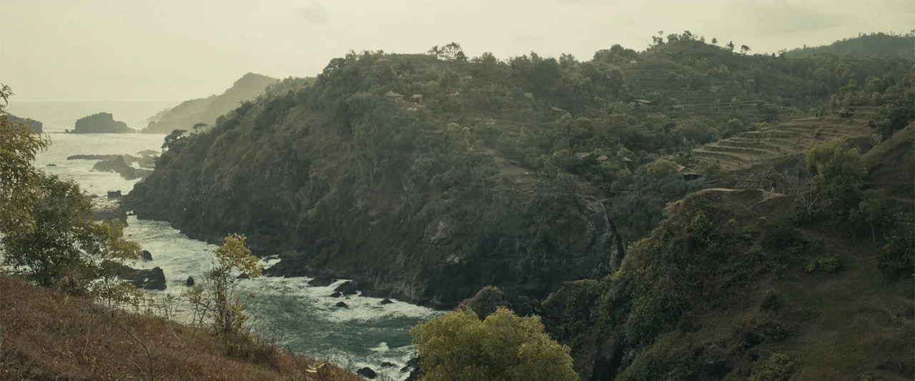 The Unholy (2022) movie still: establishing — A rugged coastline features steep rocky cliffs covered in dense tropical vegetation and trees. The…