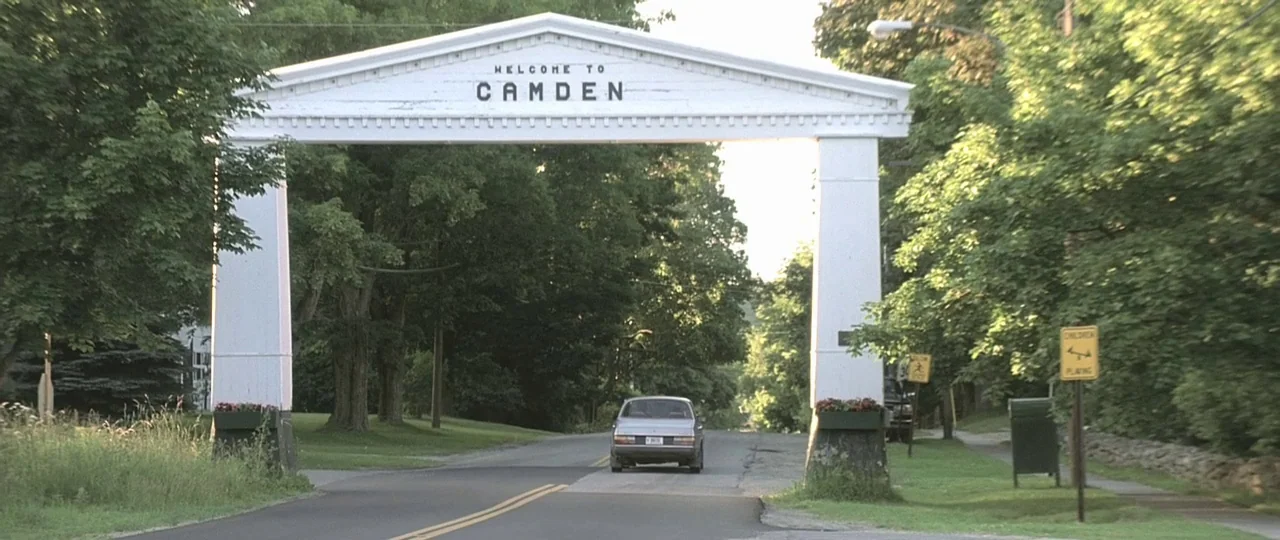 In the Bedroom (2001) movie still: wide — A white wooden archway spanning a paved road features the text 'Welcome to Camden'. A sedan drives away…