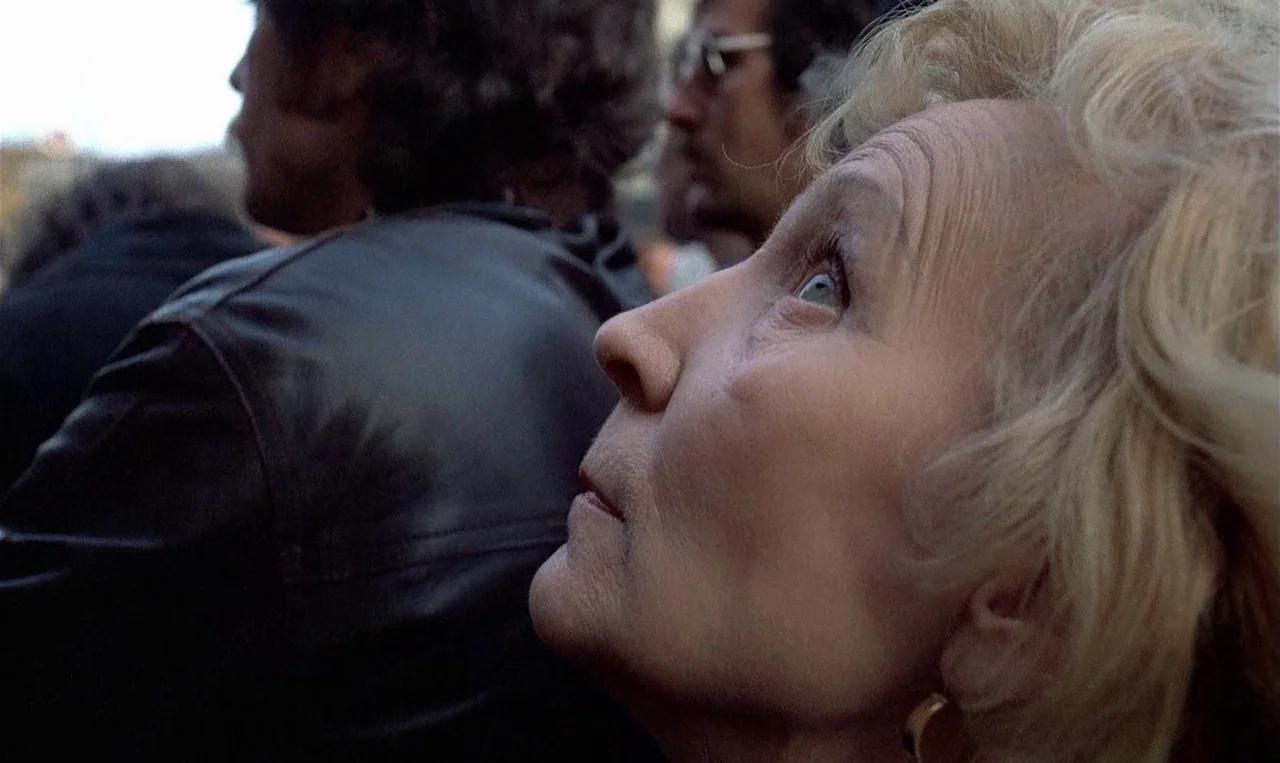 Man on the Roof (1976) movie still: closeup — An outdoor crowd scene with blurred figures of people standing in the background. An elderly woman…