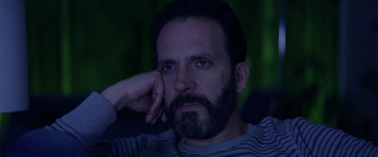 Human Animals (2020) movie still: closeup — An interior space is dimly lit by a soft blue light source. A man with a dark beard sits with his cheek…