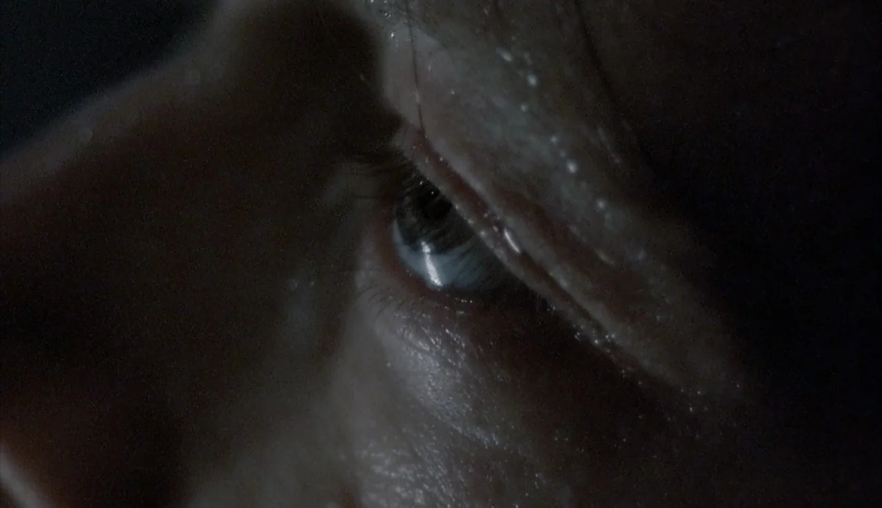 Angst (1983) movie still: closeup — An extreme close-up of a human eye captured in a dark, shadow-filled environment. The skin around the orbit…
