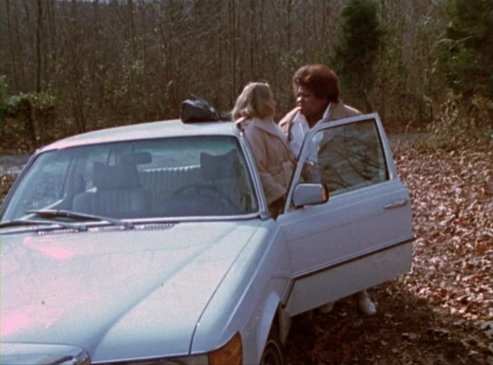 Desperate Living (1977) movie still: medium — A silver car sits parked on the side of a road bordered by dense trees. Two women stand by the open…