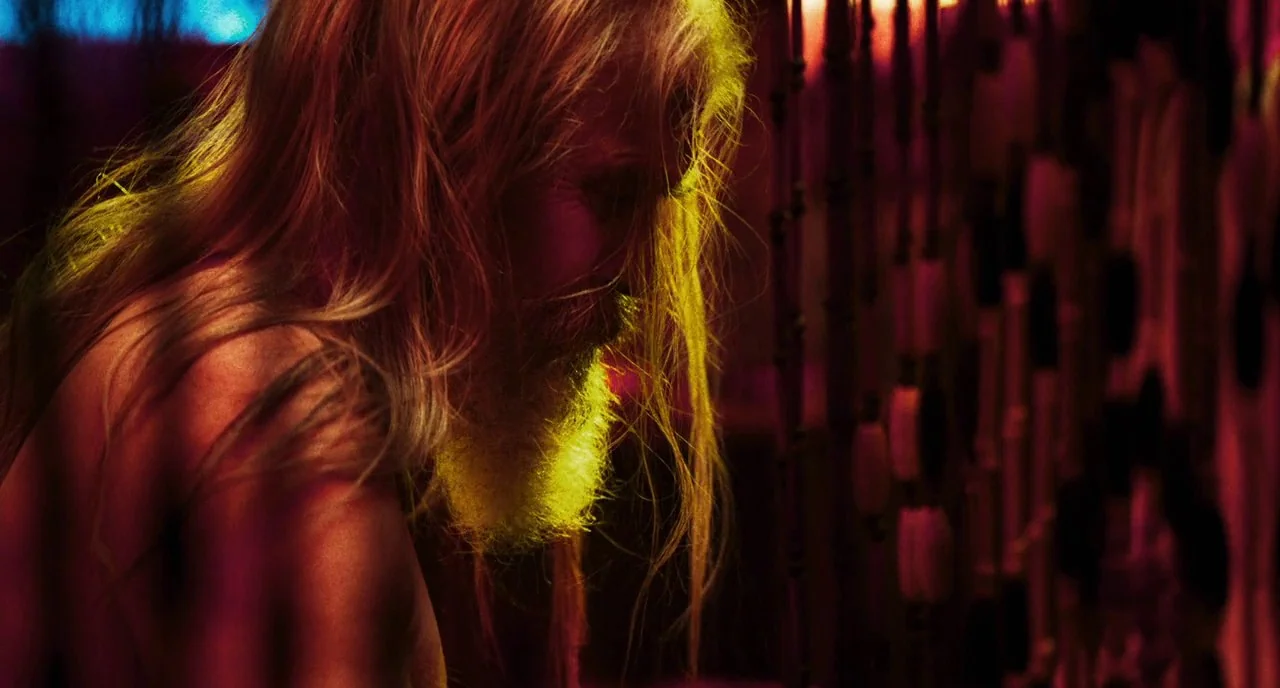 3 From Hell (2019) movie still: closeup — A dark space lit by high contrast red and yellow light. A shirtless man with long blonde hair looks down…