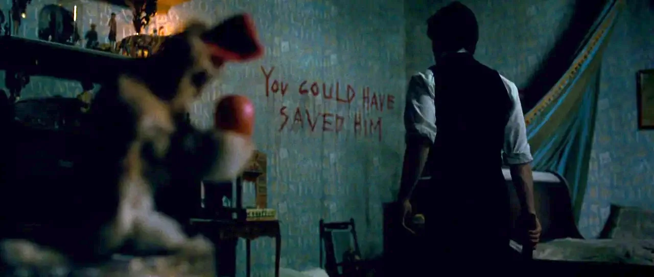 The Woman in Black (2012) movie still: medium — A dimly lit room with vintage patterned wallpaper features red text written on the wall. A man in a…
