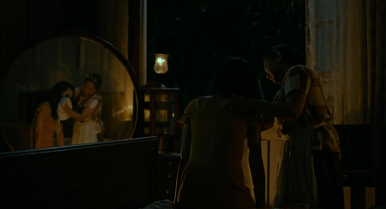 In My Mother’s Skin (2023) movie still: medium — A dimly lit room with an oval vanity mirror reflecting two women. One woman stands beside a seated…