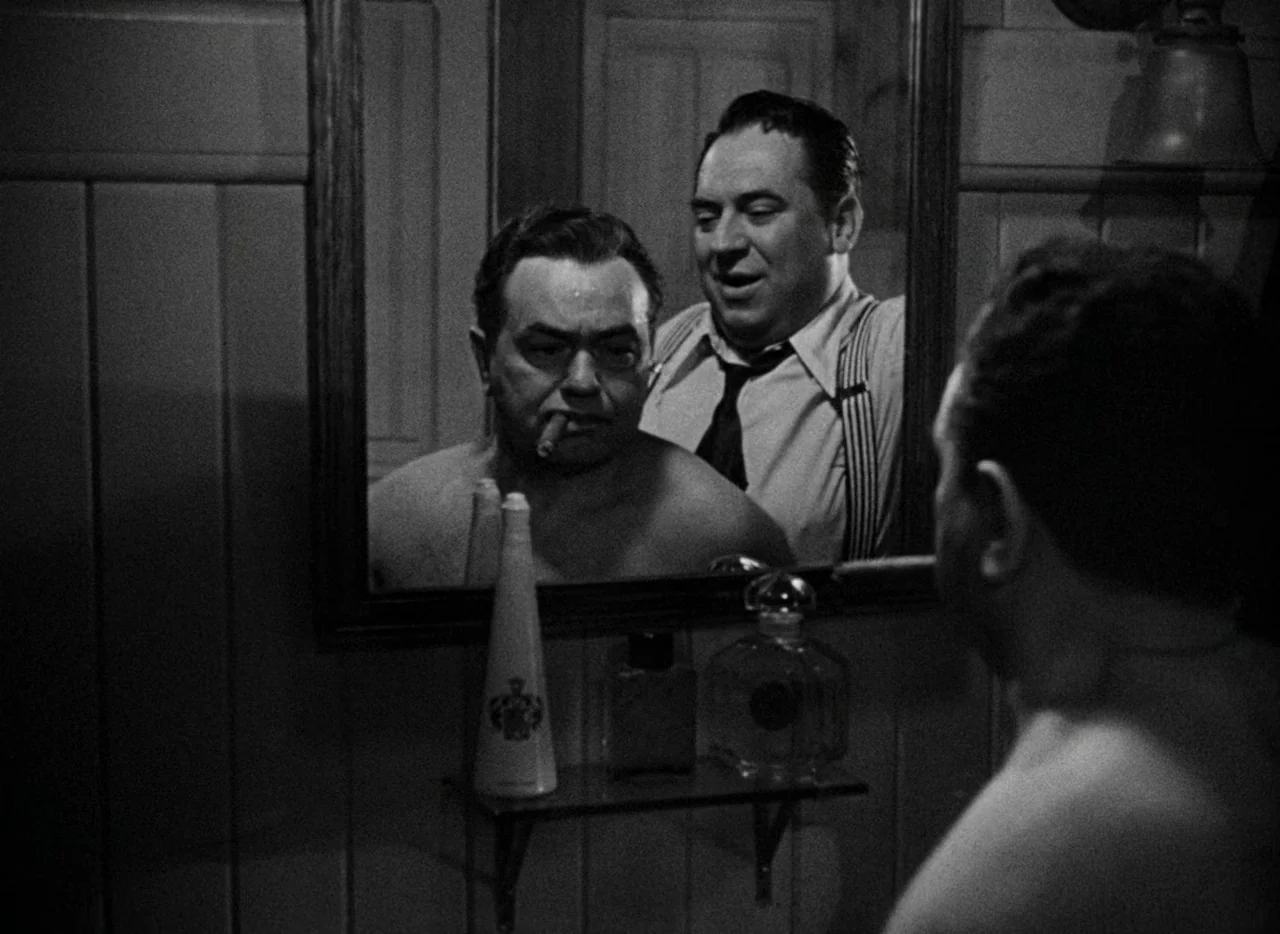 Key Largo (1948) movie still: medium — A dimly lit washroom features wooden wall paneling and a large mirror above a shelf. A shirtless man smoking…