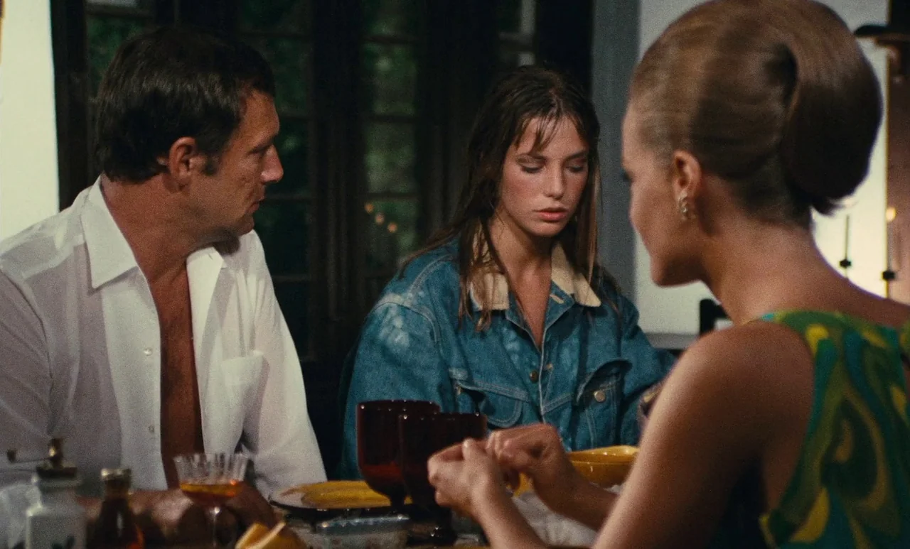 The Swimming Pool (1969) movie still: medium — A dining table with dark red glasses is set in a dimly lit room. Three individuals are seated around…