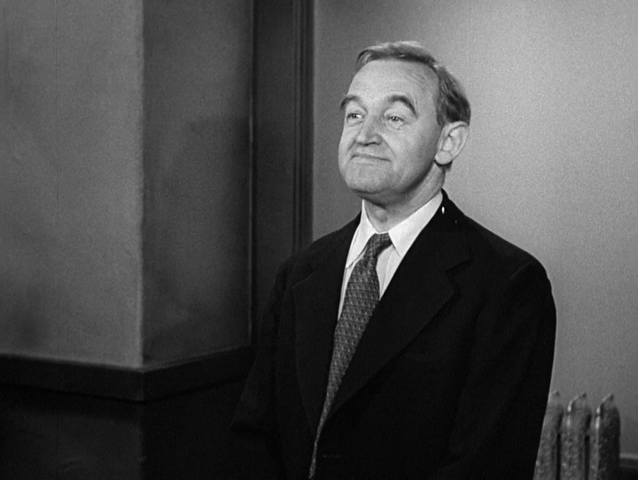 The Naked City (1948) movie still: medium — A plain interior room with a smooth wall and a vertical structural pillar. An older man in a dark suit…