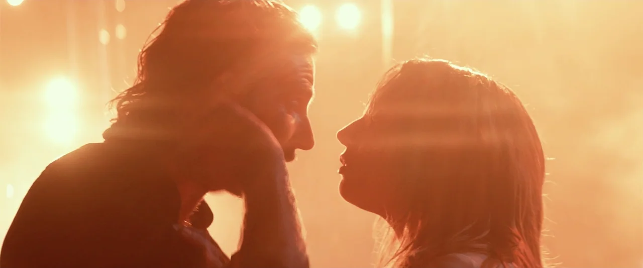 A Star Is Born (1954) movie still: medium — A dark, hazy concert stage is flooded with intense golden orange stage lighting. Two people stand facing…