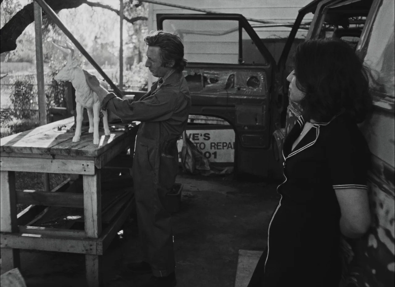 Fremont (2023) movie still: fullbody — An outdoor auto repair space with a wooden workbench and a partially dismantled van. A man examines a small…
