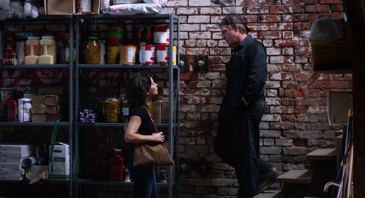 Killer Joe (2011) movie still: fullbody — A utilitarian storage area features industrial metal shelving units filled with canned goods. A man in a…