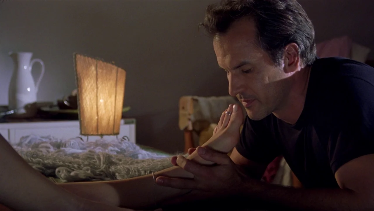 Eros (2004) movie still: medium — A dark indoor space contains a lamp and a person holding a foot. The man in the black shirt looks at the foot he…